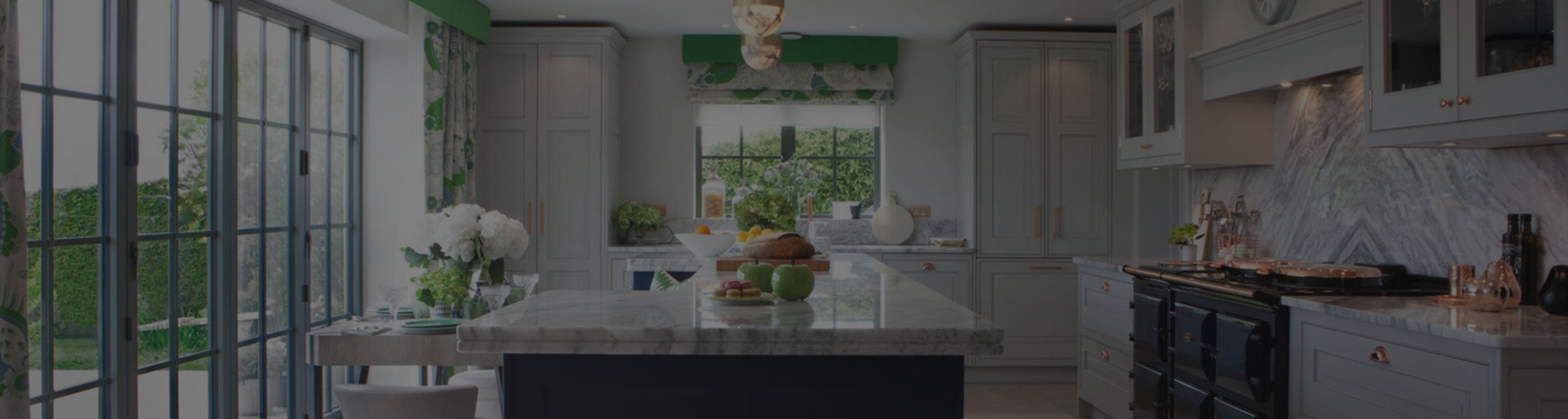 Moderne Landhausküche mit Marmor-Arbeitsplatte, Kochinsel, großem Herd, grauen Fronten, grünen Akzenten und viel Tageslicht durch bodentiefe Schiebetüren