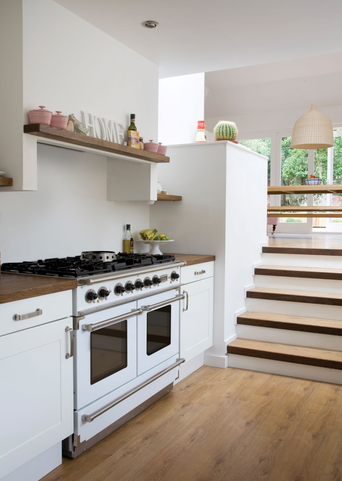 Helle, moderne Küche mit weißem Gasherd, Holzarbeitsplatte, offenem Regal mit Dekorationen und Treppe zum Essbereich mit großen Fenstern.