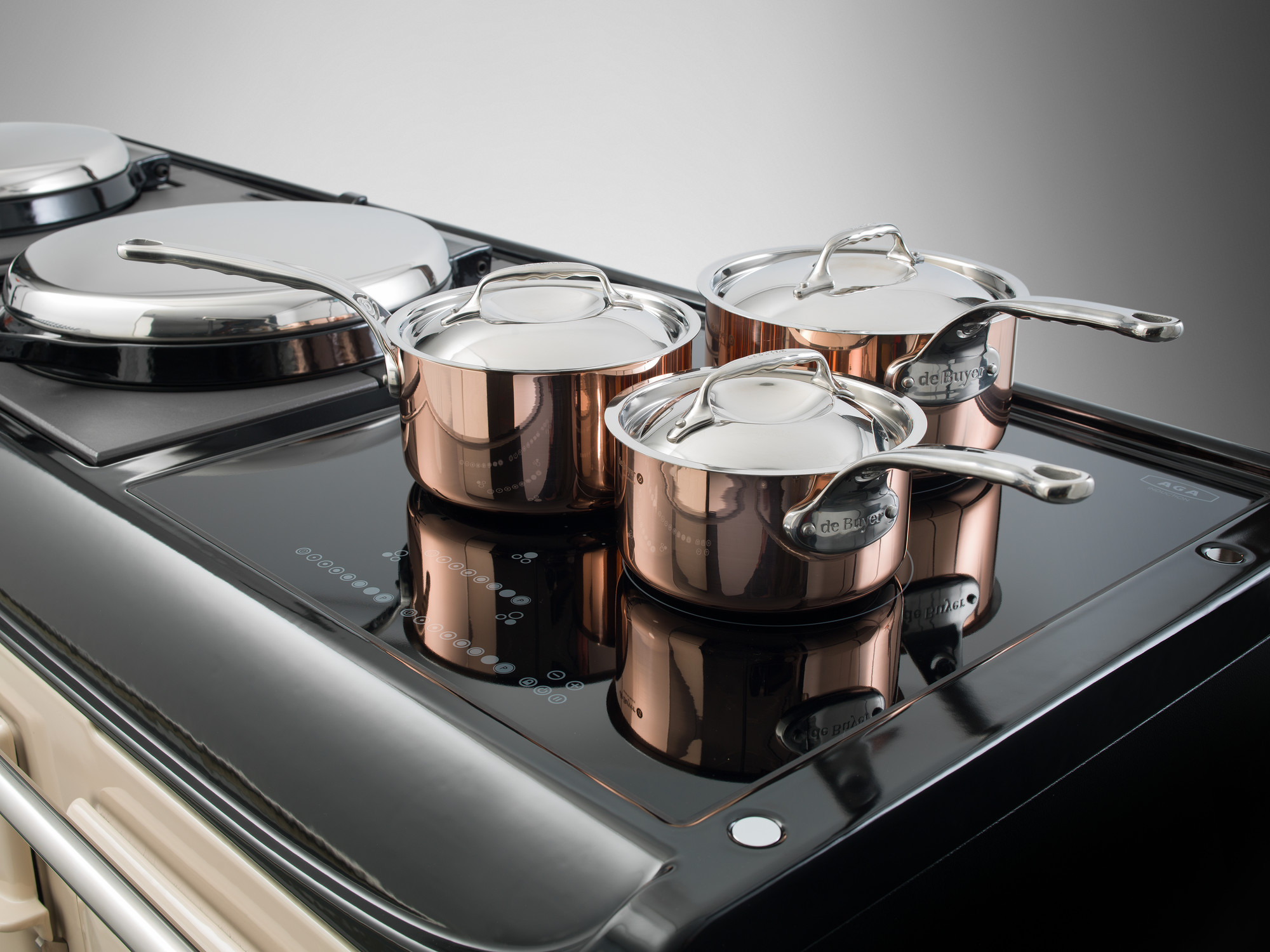 Drei glänzende Kupfer-Kochtöpfe mit Deckeln stehen auf einem schwarzen Induktionskochfeld in einer modernen Küche