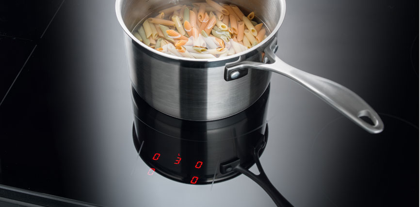 Ein silberner Kochtopf mit ungekochten, bunten Penne-Nudeln steht auf einem schwarzen Induktionskochfeld mit digitaler Anzeige.