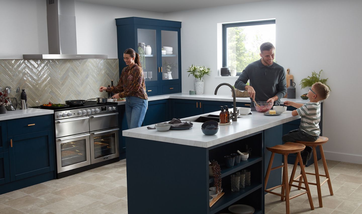 Hochwertige Einbauküche in Dunkelblau mit Kochinsel, weißer Arbeitsplatte, Herringbone-Fliesenspiegel und großem Edelstahl-Standherd; gemütliche Familienszene beim Kochen – stilvolle, funktionale Küchenplanung