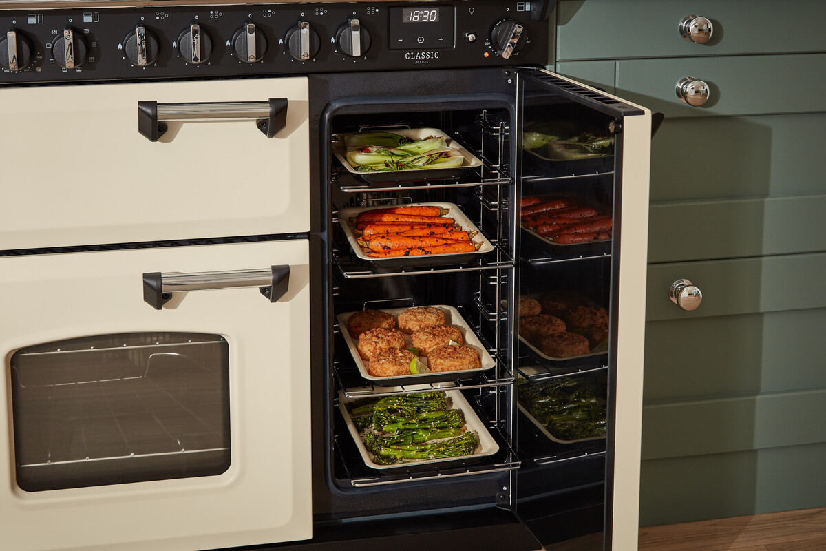 Geöffneter Range Cooker in Creme mit schwarzer Bedienblende; geräumiger Backofen mit mehreren Ebenen, auf Blechen Gemüse und Fleisch – ideal zum gleichzeitigen Garen; energieeffizienter Haushaltsofen für Familienküchen