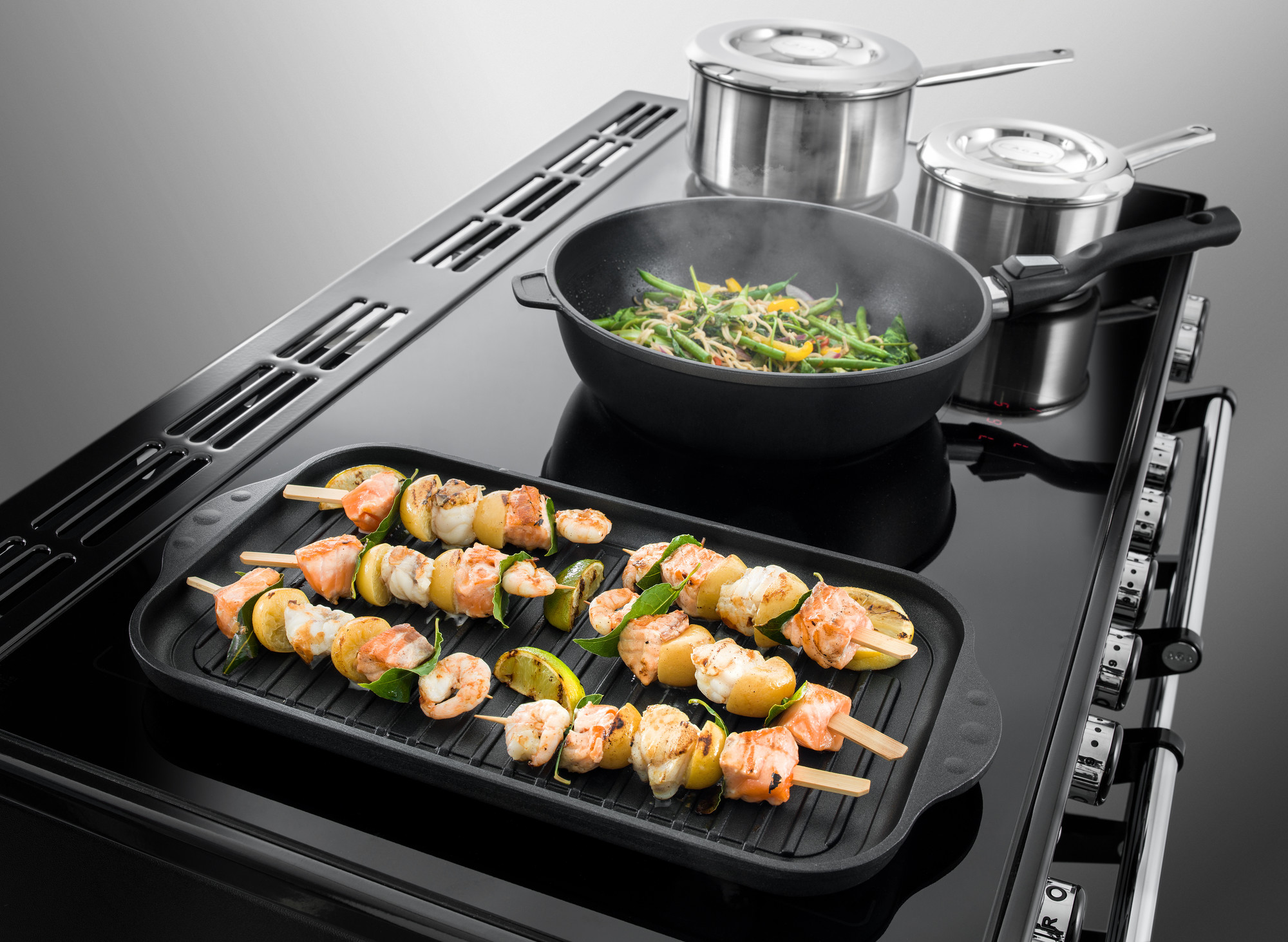 Induktionskochfeld mit Grillplatte, darauf Spieße mit Lachs, Garnelen, Kartoffeln und Gemüse, daneben Pfanne mit grünem Gemüse und zwei Edelstahltöpfe