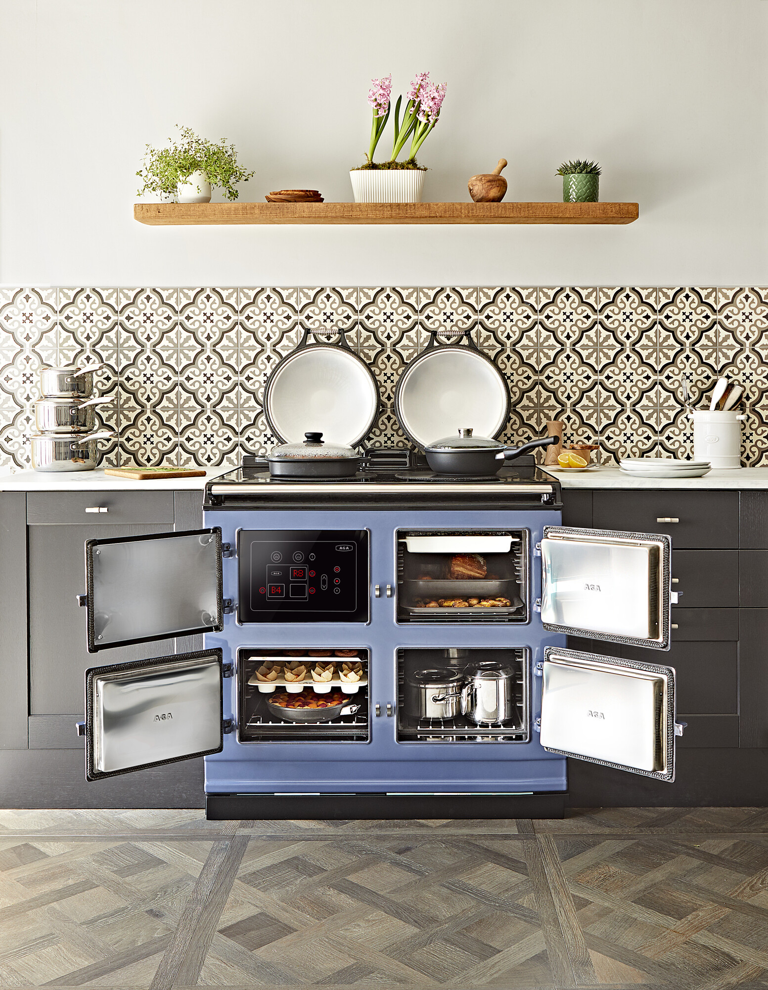 Moderner, blauer AGA-Herd mit vier geöffneten Ofentüren, darin Backwaren und Töpfe, in stilvoll eingerichteter Küche mit Blumen auf Regal und gemusterten Fliesen