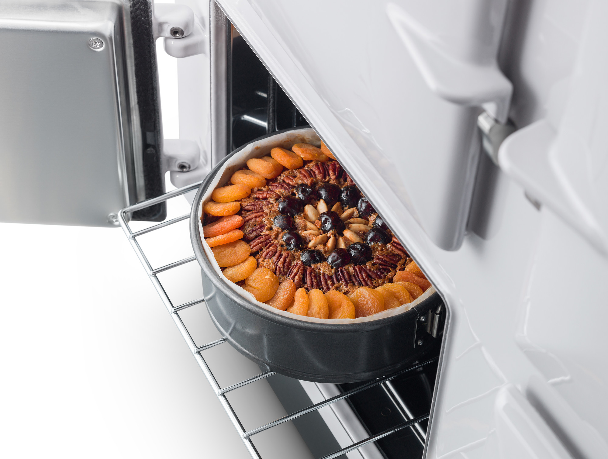 Frisch gebackener Kuchen mit Trockenfrüchten und Nüssen in einer runden Backform auf dem Backofenrost, Backofentür geöffnet