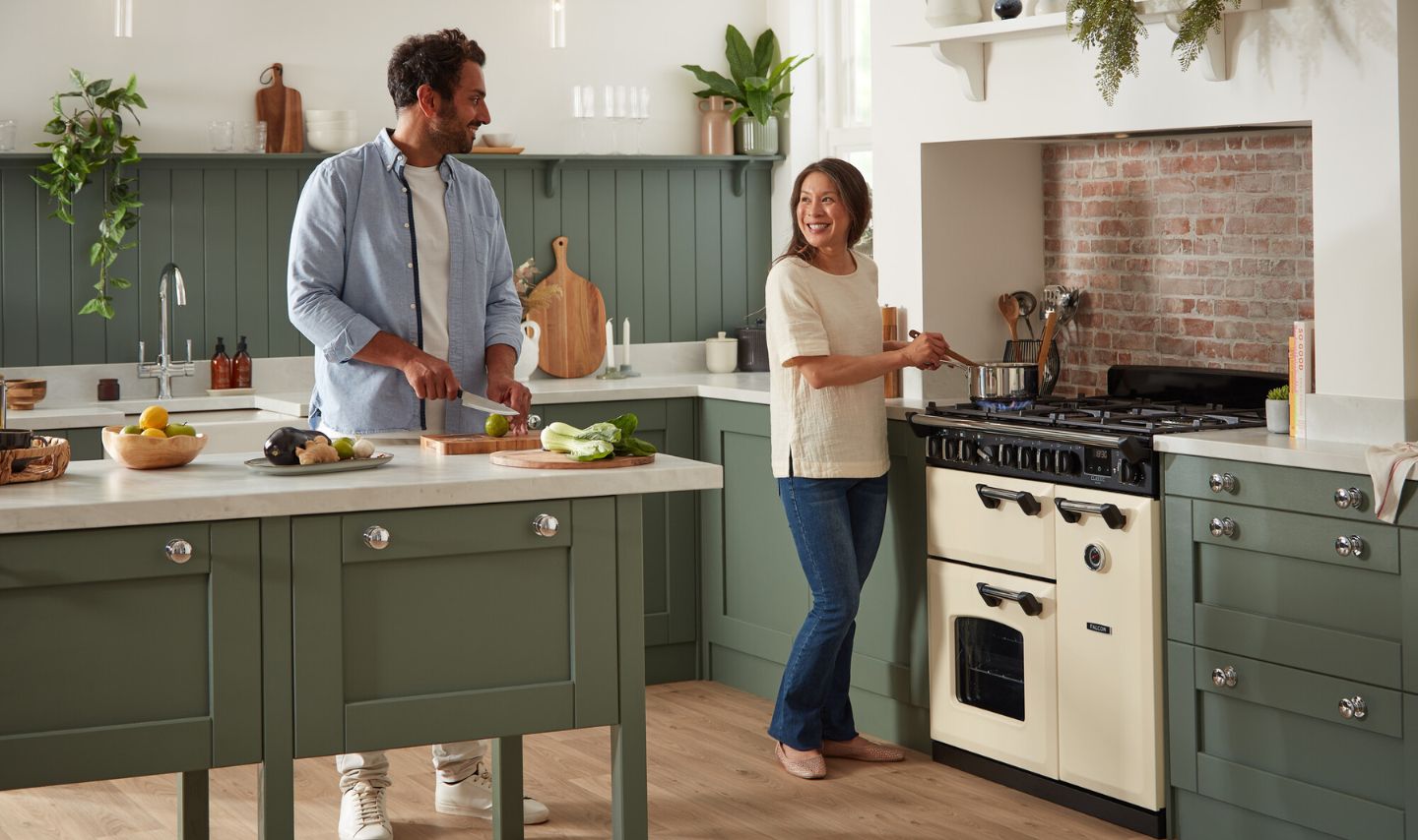 Gemütliche Küche mit grünen Shaker-Fronten, heller Arbeitsplatte, großer Kücheninsel und cremefarbenem Range Cooker; zwei Personen kochen gemeinsam, frisches Gemüse liegt bereit.