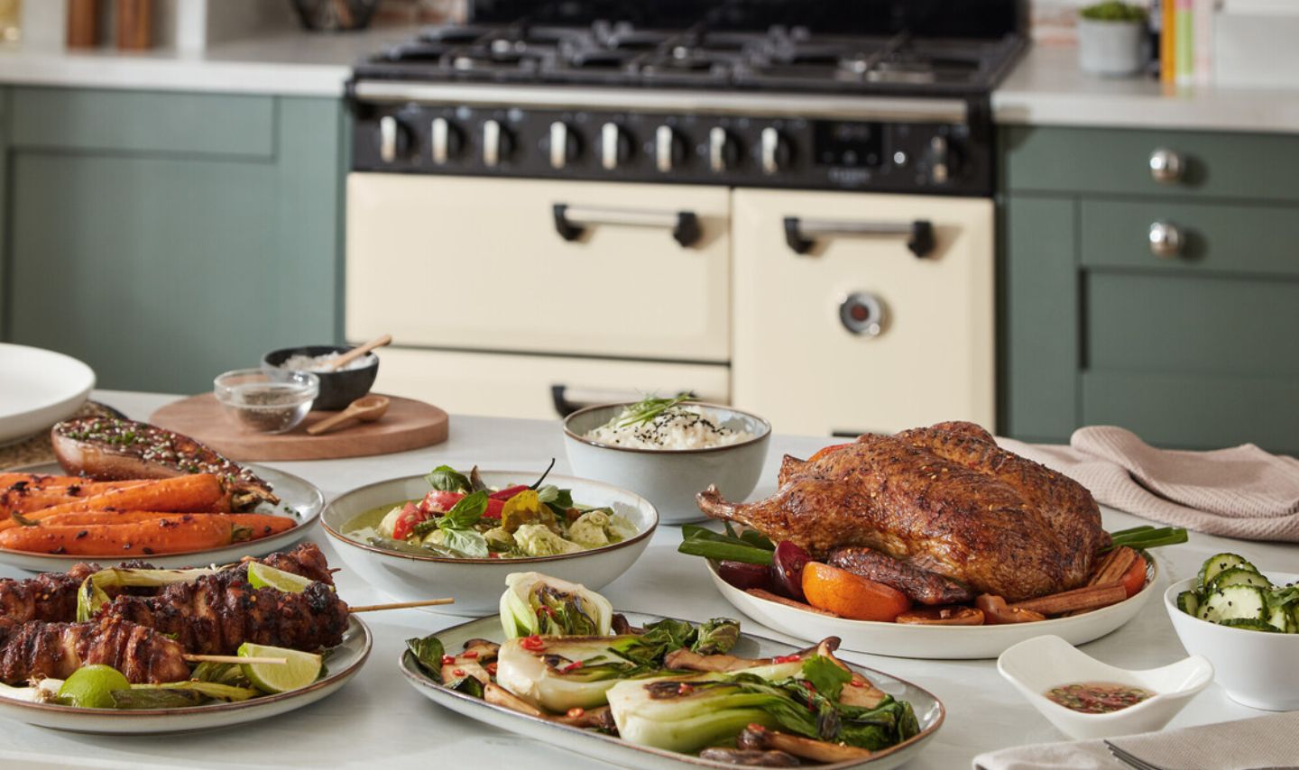 Cremefarbener freistehender Gasherd mit großem Backofen in moderner Küche; im Vordergrund servierte Speisen wie Braten, Gemüse und Spieße auf dem Tisch