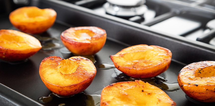Gegrillte Pfirsichhälften liegen auf einem Backblech, im Hintergrund ist ein Gasherd zu sehen