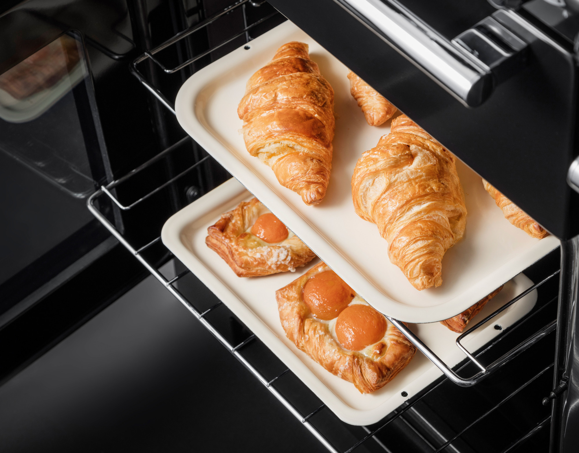Ofen mit zwei Backblechen, auf denen goldbraune Croissants und Aprikosen-Plundergebäck frisch gebacken werden