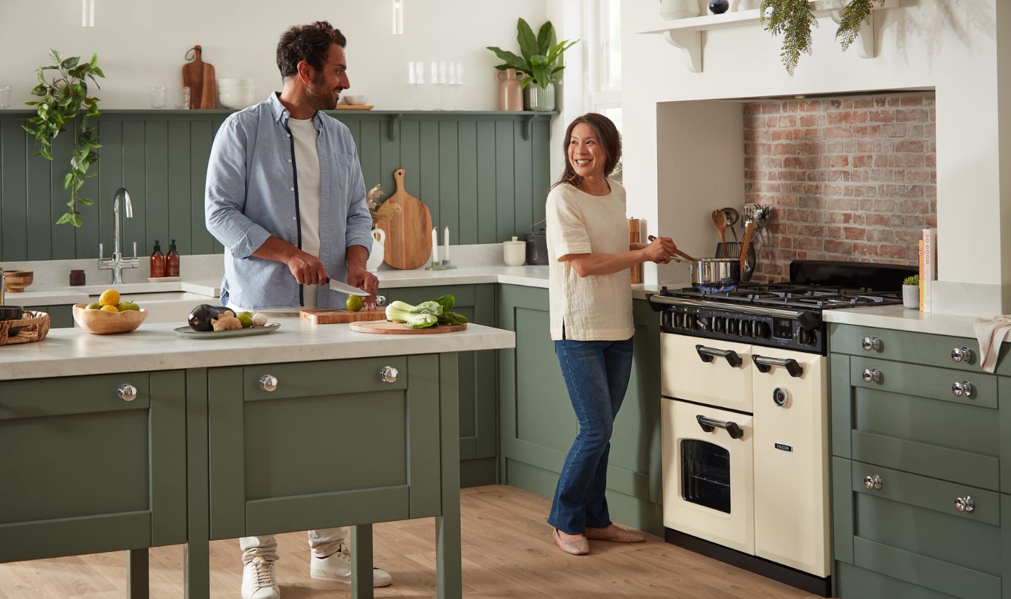Zwei Personen kochen gemeinsam in einer stilvollen Landhausküche: Er schneidet Gemüse auf der Kücheninsel, sie rührt im Topf auf einem cremefarbenen Gasherd; grüne Fronten, helle Arbeitsplatten.