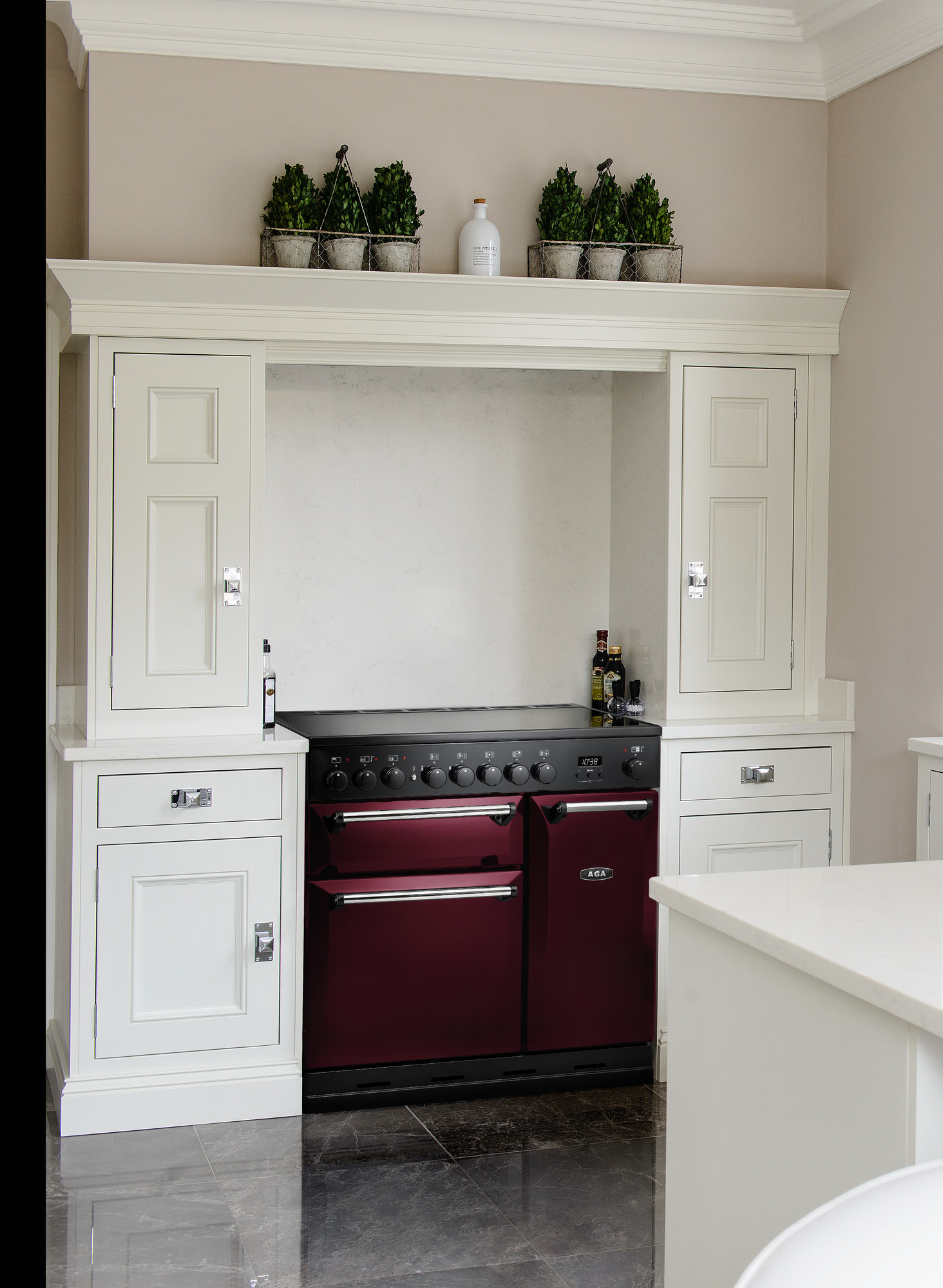 Moderner burgunderfarbener AGA-Herd mit schwarzen Bedienelementen, eingebaut in eine weiße Küche mit glänzendem Steinboden und Dekoration auf dem Küchenschrank.