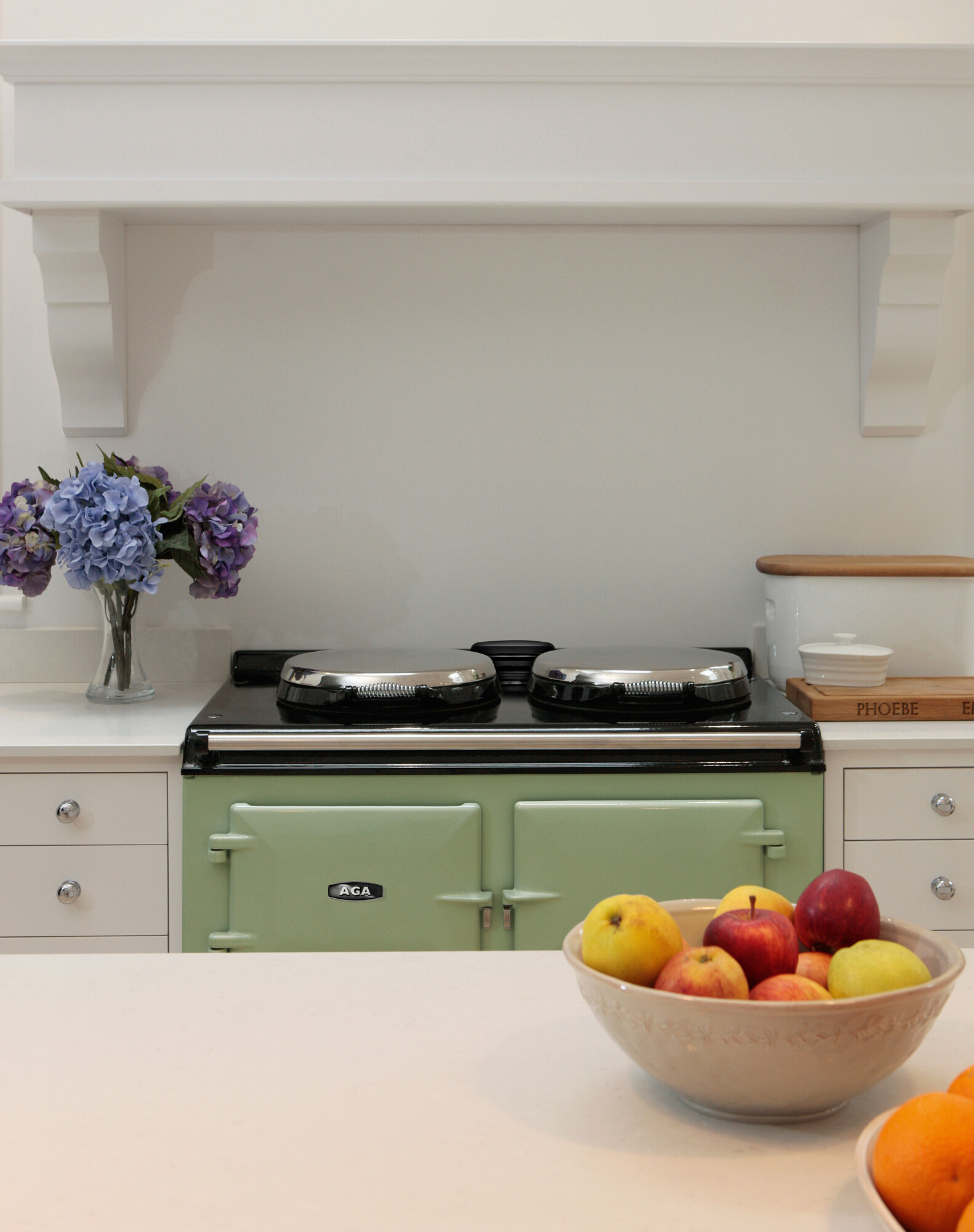 Küche mit grünem AGA-Herd, weißer Arbeitsplatte, Obstschale mit Äpfeln und Blumenstrauß aus Hortensien