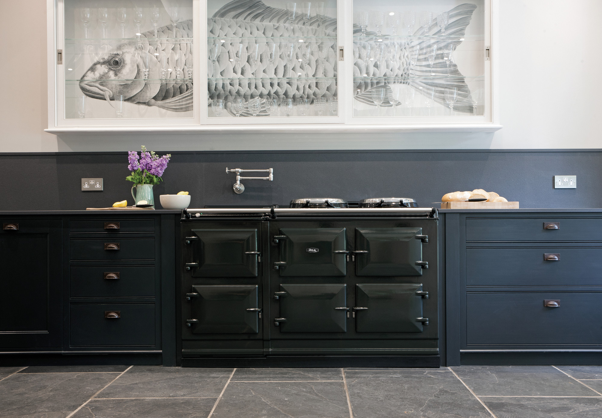 Stilvolle, moderne Küche in Schwarz mit klassischem Herd, einer Vase mit lila Blumen, Holzbrett mit Brot und auffälligem großen Fischbild hinter Glas im Oberschrank