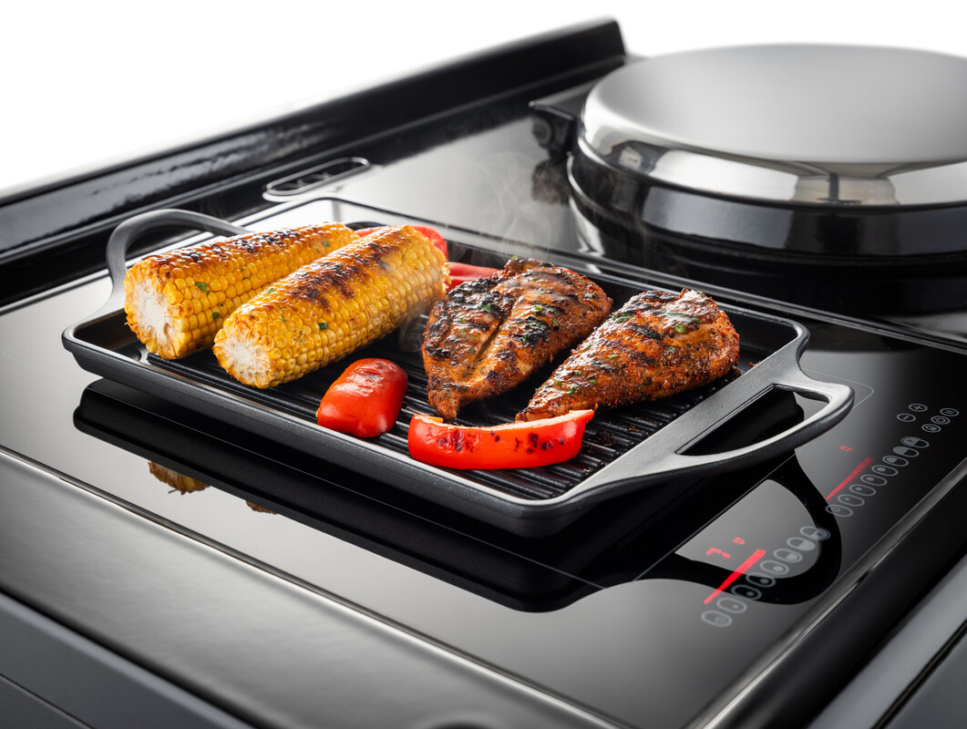 Frische Maiskolben, Fischfilets und Paprikastücke werden auf einer Grillplatte auf einem Induktionsherd gegrillt