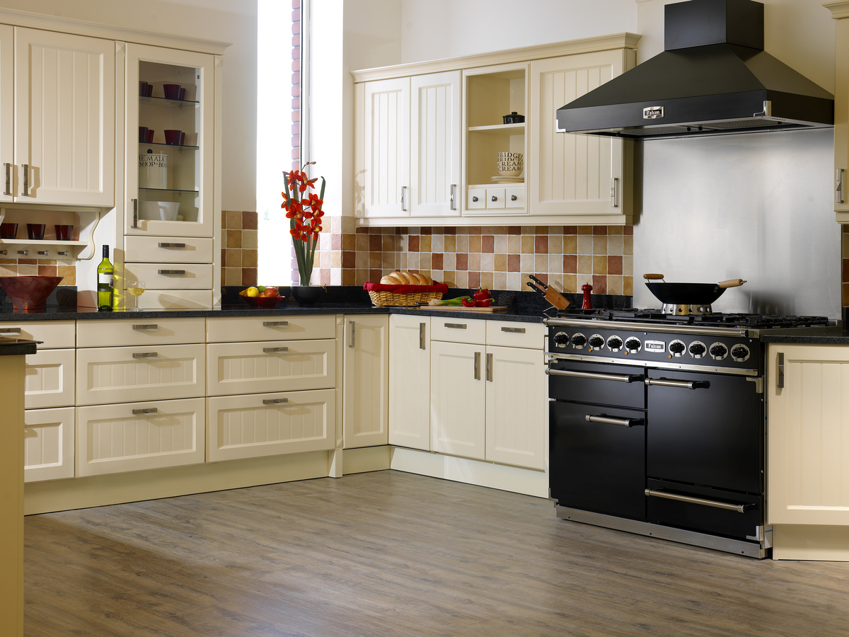 Helle Landhausküche mit cremefarbenen Schränken, großem schwarzen Gasherd, Dunstabzugshaube, Obst, Brot und Weinflasche auf der Arbeitsplatte