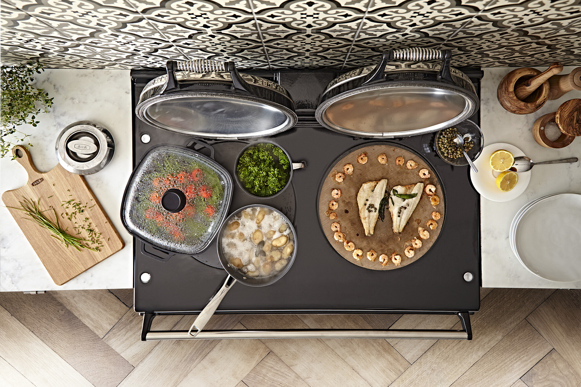 Vogelperspektive auf einen Herd mit Fischfilets, Garnelen, Kartoffeln, Gemüse und frischen Kräutern sowie modernem Kochgeschirr