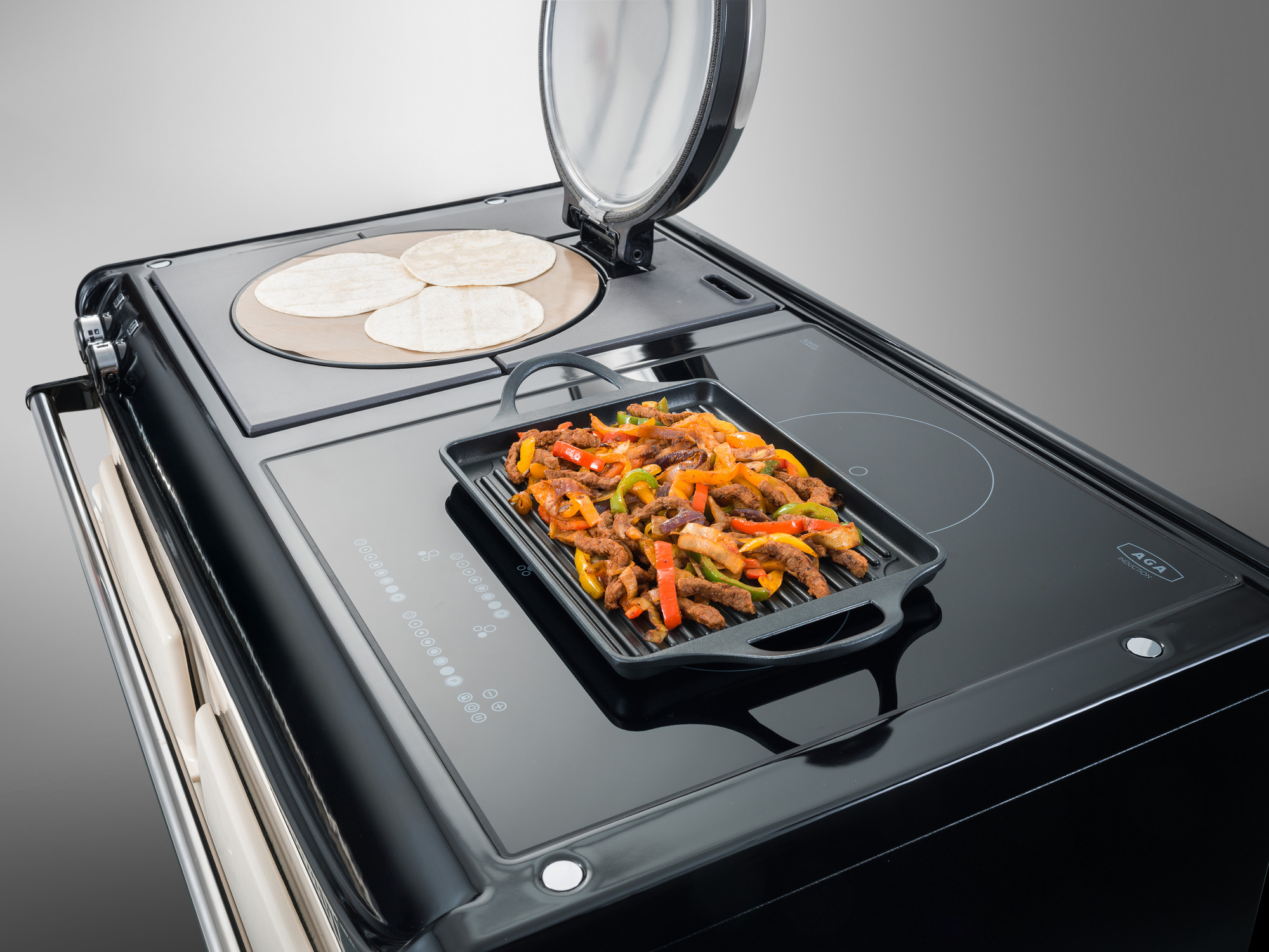 Schwarzer Herd mit Grillplatte, darauf gebratene Fleischstreifen mit Paprika, und nebenbei Tortillas auf einem separaten Kochfeld