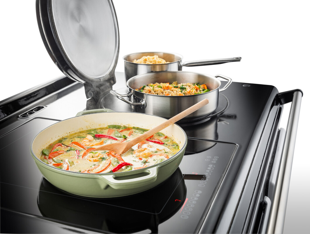 Drei Töpfe mit verschiedenen Gerichten köcheln auf einem modernen Induktionskochfeld, im Vordergrund Gemüsepfanne mit Holzlöffel