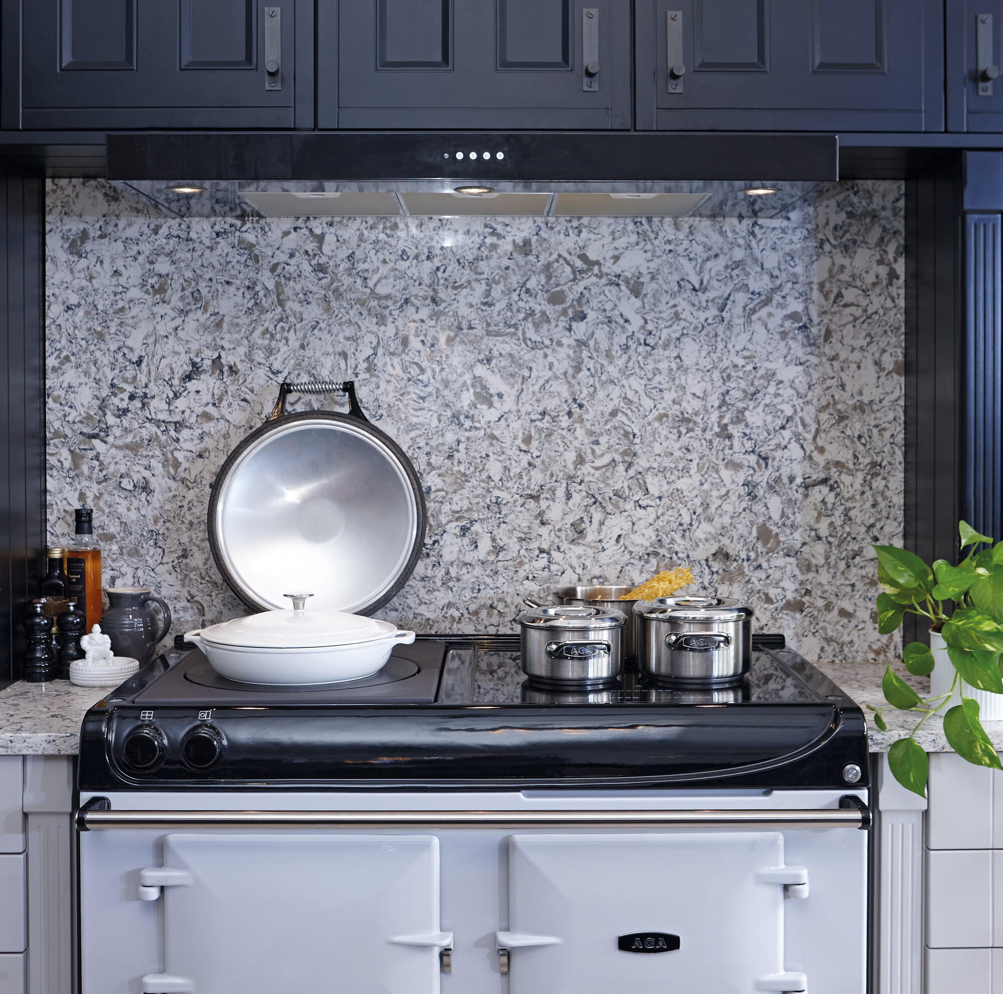 Küchenszene mit einem weißen AGA-Herd, mehreren Kochtöpfen, einer Marmor-Rückwand und Gewürzen, daneben eine grüne Pflanze